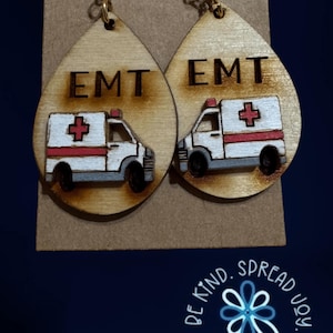 May include: Wooden teardrop earrings featuring an ambulance design with the letters "EMT" above the vehicle. The ambulance is white with red accents and a red cross. The earrings hang from gold-colored hooks.