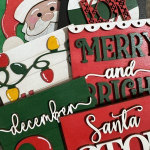 May include: A collection of Christmas-themed wooden signs and ornaments. The signs feature phrases like "Merry and Bright" and "Santa Stop Here" in white script against red and green backgrounds. A Santa Claus cutout and a "JOY" ornament are also visible.