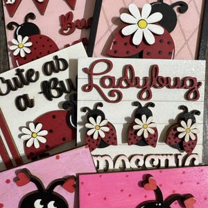 May include: A collection of wooden ladybug-themed decorations. The pieces include ladybugs with daisy accents, the word "Ladybug", and the phrase "Cute as a Bug". The color palette includes red, black, white, and pink.