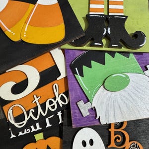 May include: A collection of Halloween-themed wooden decorations. Includes candy corn, witch legs, the word "October", a Frankenstein gnome, a ghost, and the word "Boo" with a spiderweb. The decorations are painted in orange, black, white, green, and purple.