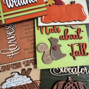 May include: A collection of wooden fall-themed signs. The signs feature phrases like "Harvest," "Nuts about Fall," and "Pumpkin Spice." Decorative elements include pumpkins, a squirrel, and autumn leaves. Colors include orange, brown, green, and white.