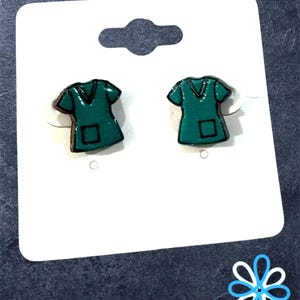 May include: A pair of teal-colored scrub top earrings with black outlines, displayed on a white card with a hang hole. The earrings are shaped like miniature medical scrubs. A blue and white floral design is in the bottom right corner.