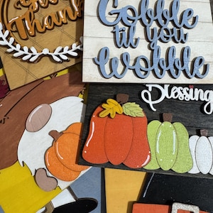May include: Wooden fall-themed decorations with phrases like "Give Thanks" and "Gobble til you Wobble." The image includes pumpkins, a gnome, and a pilgrim hat. The decorations are in various colors, including orange, green, and brown.