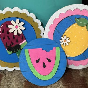 May include: Three wooden coasters with summer fruit designs. One features a watermelon popsicle, another a lemon, and the third a watermelon slice. Each coaster has a scalloped edge and a daisy accent, perfect for a kitchen or dining room.