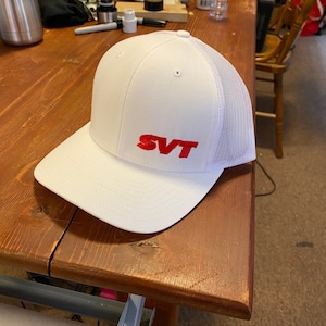 May include: White baseball cap with a red embroidered SVT logo on the front. The cap has a mesh back.