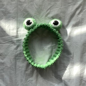 Op de afbeelding: Groen gehaakt hoofdband met twee grote, ronde, witte en zwarte ogen. Het hoofdband is ontworpen om eruit te zien als een kikker.