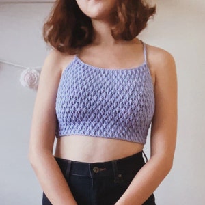 Könnte beinhalten: Ein lilafarbenes Häkel-Crop-Top mit einem zarten, strukturierten Muster. Das Top hat dünne Träger und eine figurbetonte, verkürzte Silhouette.