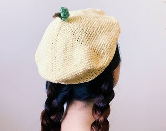 May include: A yellow crocheted beret with a green leaf detail. The beret is worn by a person with long brown braids.