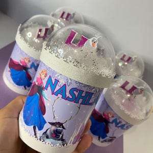 May include: Several personalized snow globe tumblers featuring characters from the movie Frozen. The tumblers have a white base with a purple and blue design, the name "NASHI" and the letter "L". The clear dome tops are filled with glitter and small figures.