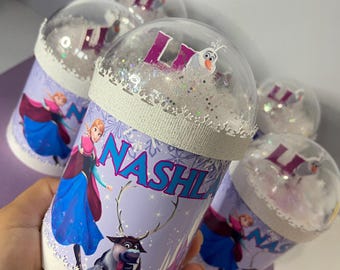 May include: Several personalized snow globe tumblers featuring characters from the movie Frozen. The tumblers have a white base with a purple and blue design, the name "NASHI" and the letter "L". The clear dome tops are filled with glitter and small figures.
