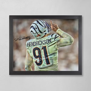 May include: Framed print featuring a football player in a jersey with the name "Hendrickson" and the number "91". The player is wearing a helmet with a black and white design and is raising their index finger. The print is in a black frame.
