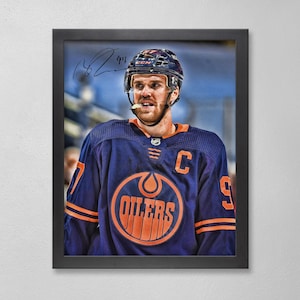 May include: Framed portrait of a hockey player in an Oilers jersey. The jersey is navy blue with orange accents and the Oilers logo. The player wears a helmet and has a signature in the upper left corner. The frame is black.