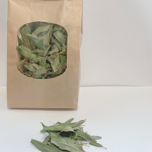 Puede incluir: Una bolsa de papel marrón con una ventana transparente que muestra hojas verdes secas en su interior. Un montón de hojas verdes secas se encuentra sobre una superficie blanca.