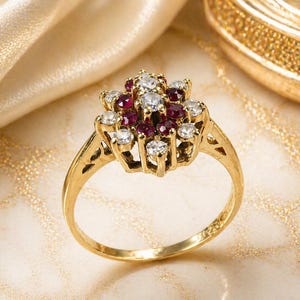 May include: A gold ring with a cluster of gemstones. The ring's centrepiece features a floral arrangement of round-cut, clear diamonds and deep red rubies. The band is polished gold, set against a soft, golden background.