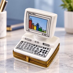 May include: A white porcelain trinket box shaped like a vintage desktop computer. The screen displays a colorful bar graph with numerical data. The keyboard is detailed with black lines. The base features a metallic gold-toned hinged closure.