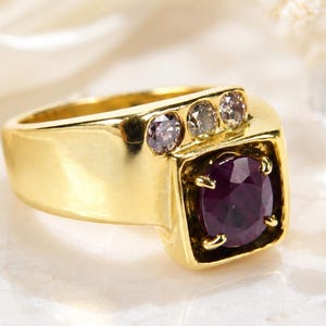 May include: A gold ring featuring a large, round, deep purple gemstone set in a square-shaped bezel. The band includes three small, round, light-colored accent stones arranged in a row along the side of the central setting.