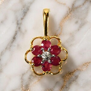 May include: A gold pendant in the shape of a flower, featuring six deep red gemstones arranged around a central, smaller, clear gemstone. The pendant has a loop at the top for a chain.