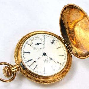 May include: A gold pocket watch with an open face and a white dial. The watch has Roman numeral hour markers and a small sub-dial for seconds. The watch is engraved with the name "Crescent" and the words "15 Jewels Adjusted."