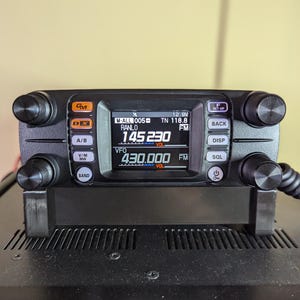 May include: A black handheld radio transceiver with a digital display showing "145.230 FM" and "430.000 FM". The radio has several buttons and knobs, including a volume knob, a band knob, and a squelch knob.