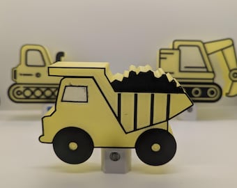 May include: A yellow and black dump truck night light with a black load in the bed. The night light has black wheels and a white window. Other construction vehicles are in the background.