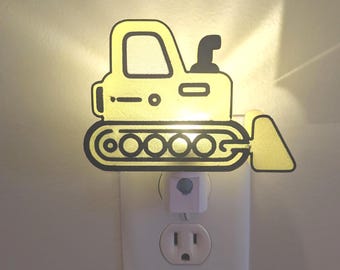 May include: A yellow and black bulldozer-shaped night light. It is plugged into a white wall socket. The light shines from the front of the bulldozer.