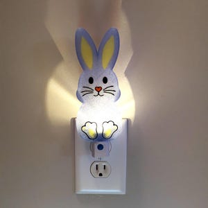 May include: A blue bunny night light plugged into a standard electrical outlet. The bunny has yellow ears and a white face with black whiskers.