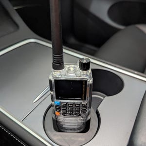 May include: A clear, black and orange two-way radio with a long antenna is in a cup holder in a car. The radio has a digital display and a keypad with numbers and symbols. The radio is labelled "Tigradio".