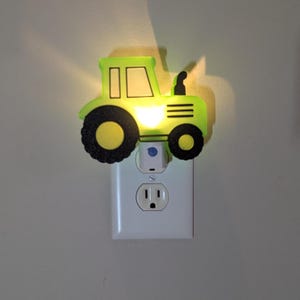 May include: A green tractor-shaped night light plugged into a white electrical outlet. The tractor has black wheels and a yellow center, with a light shining from the cab. The night light is designed to provide a soft glow in a child's room.