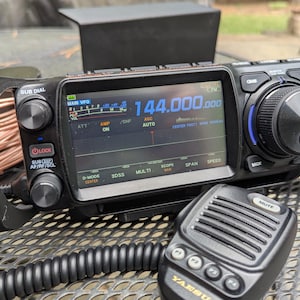 May include: A black and gray Yaesu amateur radio transceiver with a digital display showing 144.000.000. The radio has multiple dials, buttons, and a connected microphone. The microphone has the text "MUTE" on it.