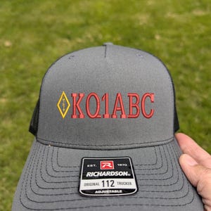 May include: Gray Richardson 112 trucker hat with a black mesh back. The front of the hat features the text "K01ABC" in red embroidery, with a yellow and red diamond design. The hat has an adjustable strap.