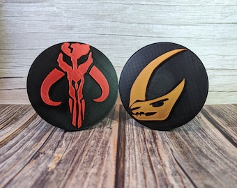 May include: Two round black coasters with a red and white Mandalorian symbol on one and a gold and white symbol on the other.