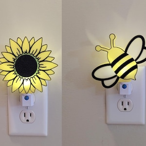 May include: Two night lights plugged into electrical outlets. The first night light is shaped like a sunflower with yellow petals and a black center. The second night light is shaped like a bee with yellow and black stripes.