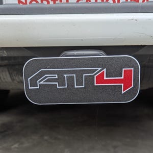 May include: A black and gray trailer hitch cover with the letters "AT4" in white and red. The cover is rectangular with rounded corners and is mounted on the back of a vehicle.