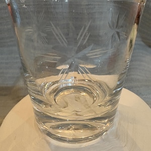 May include: A clear glass tumbler with a starburst pattern etched into the side. The glass is sitting on a white surface.