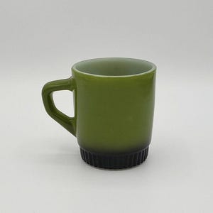 Puede incluir: Taza verde oliva con base negra. La taza tiene un asa redondeada y forma cilíndrica. La base tiene un diseño acanalado. El interior de la taza es de color verde claro. La taza es de cerámica.