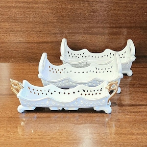 May include: Three white porcelain rectangular dishes with scalloped edges and intricate lattice cutouts. Each dish features gold-painted handles and trim, resting on four small feet. The pieces are arranged in a staggered formation on a surface.