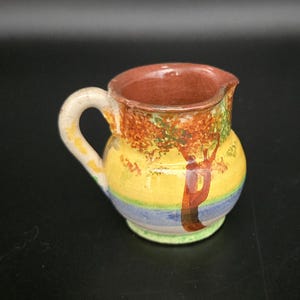 May include: A small, hand-painted ceramic pitcher with a rounded body and a handle. The pitcher features a landscape design with a yellow background, a tree, and blue and green accents. The interior is brown.