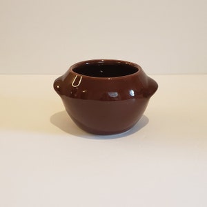 May include: A small, brown ceramic pot with rounded sides and two small handles. The pot is glazed and has a smooth, shiny finish.