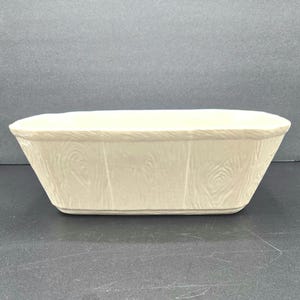 May include: A cream-coloured, rectangular ceramic bowl with a textured wood grain pattern. The bowl has a slightly flared shape and a smooth rim. The design is simple and rustic, suitable for serving or display.