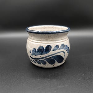 May include: A small, round ceramic pot with a speckled, off-white base. The pot features a dark blue rim and a stylized leaf pattern in dark blue around the body. The pot is likely used for decorative purposes.