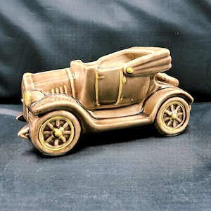 Pode incluir: Um vaso de plantas de carro vintage de cerâmica marrom com detalhes amarelos nas rodas e acabamentos. O carro conversível tem um design detalhado, incluindo faróis e uma capota conversível. O vaso é projetado para conter pequenas plantas ou flores.