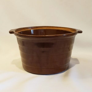 Puede incluir: Una olla de cerámica marrón con dos asas. La olla tiene un acabado esmaltado y está diseñada con un patrón acanalado.