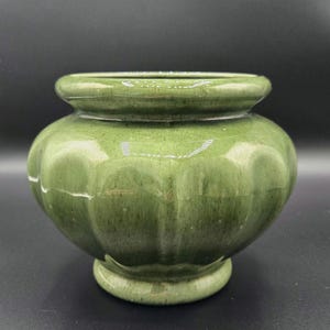 May include: A green ceramic vase with a rounded body and a flared rim. The vase has a glossy finish and a ribbed texture. The base is a smaller, rounded pedestal. The vase is likely for decorative purposes.