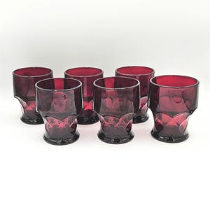 May include: A set of six vintage ruby red drinking glasses. Each glass has a decorative base and a cylindrical top. The glasses are made of a deep red glass and are arranged on a white background.