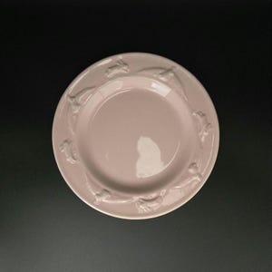 May include: A pale pink ceramic plate with a raised border featuring a repeating rabbit design. The plate has a smooth, glossy finish and a slightly recessed center. The plate is set against a dark background.