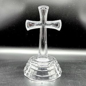 May include: Clear glass cross with a detailed design. The cross is mounted on a tiered, faceted base. The transparent material allows light to pass through, creating a reflective effect. The cross is a religious symbol.