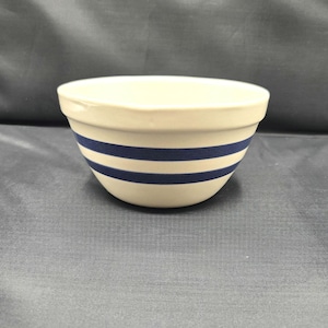 May include: Cream-colored ceramic mixing bowl with two navy blue stripes. The bowl has a rounded shape and a smooth, glossy finish. A classic kitchenware item.