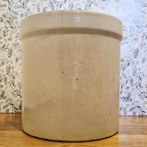 Puede incluir: Un tarro cilíndrico de gres beige con una banda ligeramente más oscura cerca de la parte superior. El recipiente de estilo antiguo tiene un acabado liso y brillante y se coloca sobre un fondo de papel pintado con motivos de hojas y una superficie de madera.