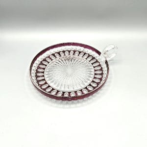 Puede incluir: Un plato de vidrio transparente con un borde morado y una pequeña asa. El plato tiene un patrón decorativo de pequeños círculos alrededor del borde.