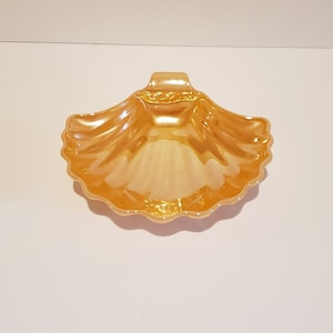 May include: A gold-colored ceramic seashell dish with a handle. The dish is designed to look like a seashell and has a smooth, glossy finish.